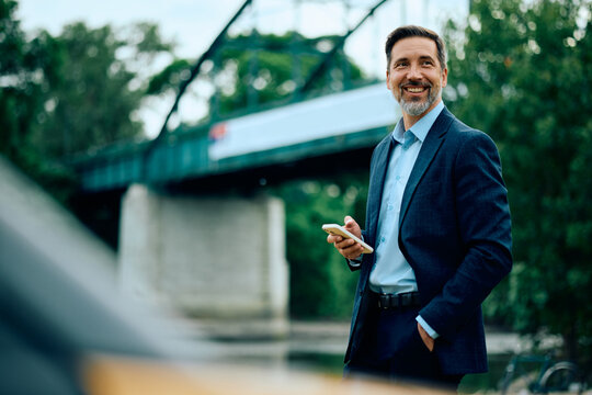 Happy businessman holding cell phone while standing outdoors and looking away.