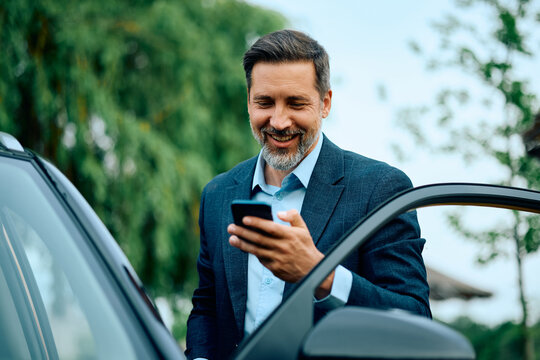 Happy businessman using smart phone while getting into his car.