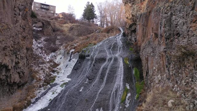 An aerial drone shot captures the majestic Jermuk Waterfall, known as Mermaid's Hair, cascading down rocky cliffs amidst lush green vegetation in Armenia's beautiful mountain landscape during spring.
