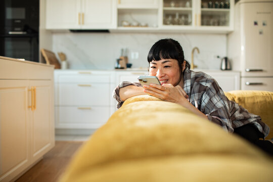 Smiling woman texting on smartphone on sofa in home kitchen