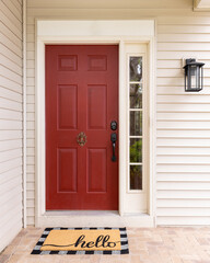 A front door detail with brown vinyl siding, a red front door, black mounted light, and a welcome may that says 