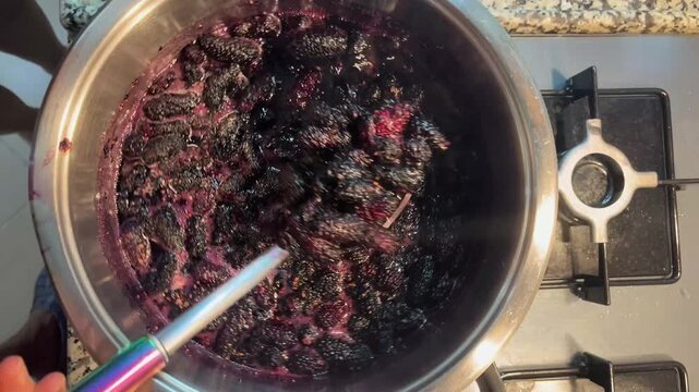 Fresh black mulberry jam is being stirred with a spoon as it simmers in a pot. Traditional homemade jam-making.