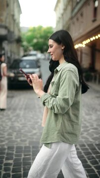 A happy young brunette woman walks down a European cobblestone street, smiling as she scrolls through social media or texts on her smartphone. Vertical Shot
