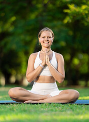 Obraz premium Young woman yoga teacher performs carry out exercises in solitude outdoors, in shady city park alone. She does exercises and repeats lotus position, padmasana