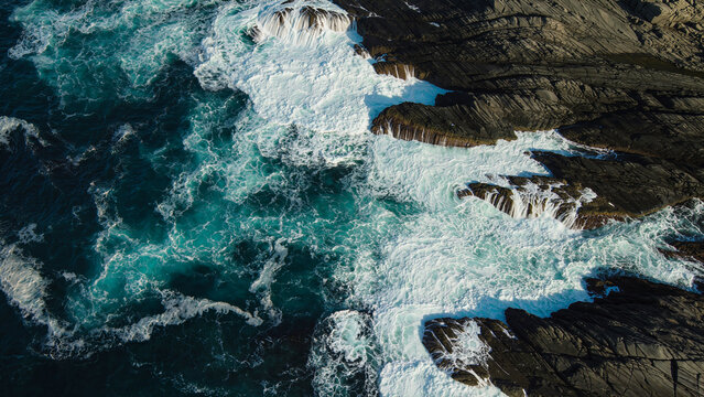 Top down aerial view of powerful turquoise ocean waves crashing against dark coastal rocks and forming white foam, concept of wild sea elements