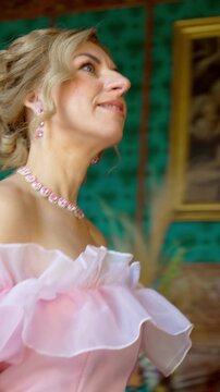 ​Close-up shot of a beautiful mature blonde woman showcasing elegant crystal jewelry, pink dress, and a sophisticated hairstyle in a luxury interior. Vertical 9:16 format.
