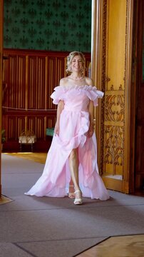 ​Beautiful blonde mid-adult woman in a luxury pink evening dress walking through an opulent palace room. Confident smiling model in a gown with a high slit. Vertical 9:16 format.