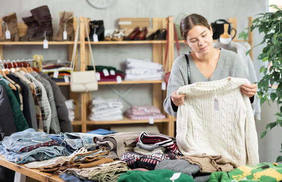 Young female shopper choosing knitted sweater in clothing store