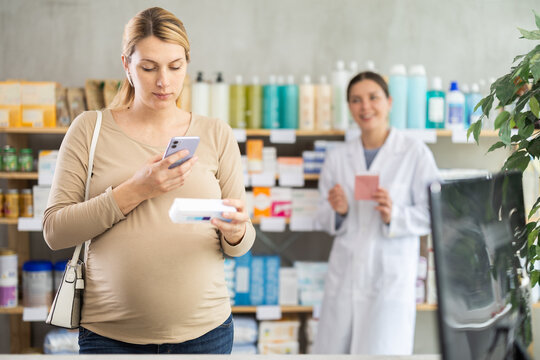 Pregnant woman in pharmacy scans QR code on diaper rash ointment product, checks certificate on manufacturers website. Girl pharmacist offer similar product in background.