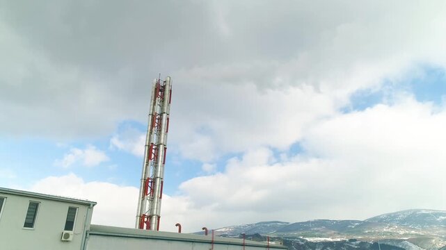 Aerial drone shot ascending past a tall smoking chimney at a district heating plant in Pirot Serbia, revealing roads, fields, lakes and snowy mountains.