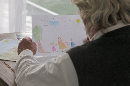 Hands of an ederly man sitting at home looking at children drawings, expressing sadness, loneliness, nostalgia, and memories of family and childhood.