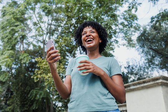 young african american woman using mobile phone and holding coffee cup in spain city in Europe at summer, black female with afro hair