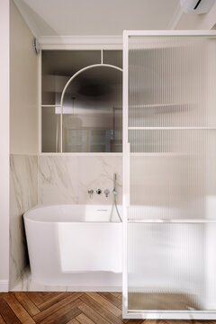 White freestanding bathtub in modern bathroom with marble and ribbed glass