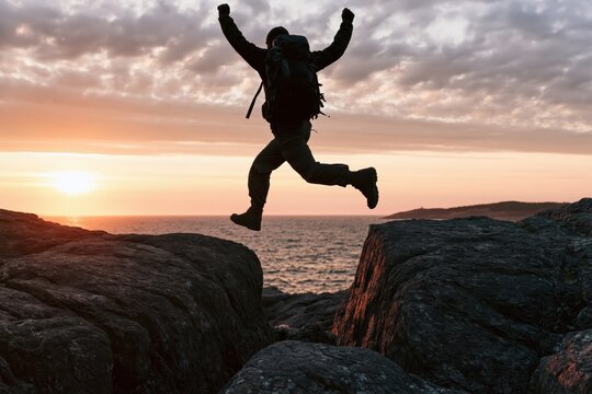 Hiker silhouette jumping over a rock gap at sunset for adventure and travel success