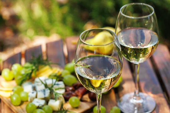 White wine glasses with rosemary garnish, cheese board and grapes on wooden table, sunny outdoor picnic, fresh gourmet pairing and relaxed lifestyle