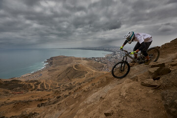 Fototapeta premium A seasoned athlete drops into a raw rocky descent at El Morro Solar, with the entire city of Lima and the Pacific Ocean laid out below, a backdrop that turns every run into something cinematic.
