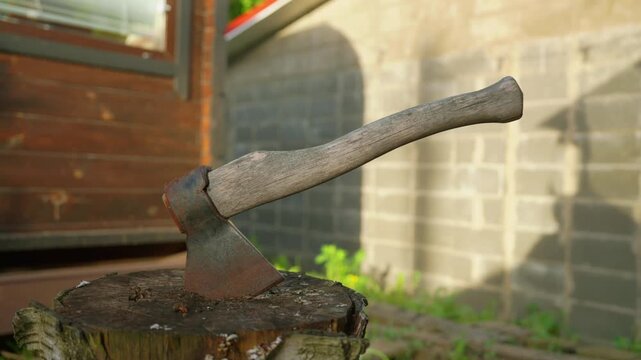 Rusty old axe with a weathered wooden handle firmly stuck in a tree stump outdoors. A traditional lumberjack tool for chopping firewood, set against a rustic backdrop in the summer sun