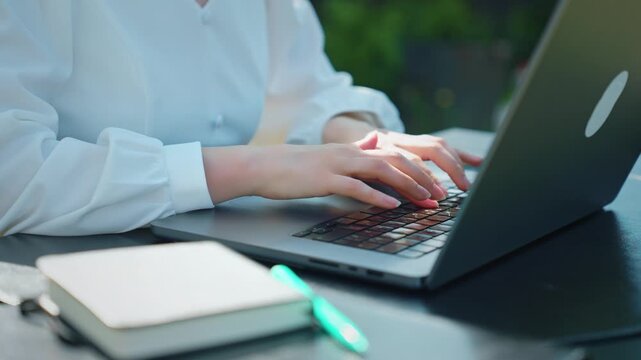 Close up of female hands typing on a laptop keyboard, working remotely at an outdoor cafe table with a notebook nearby, showcasing a modern business and technology concept on a sunny day