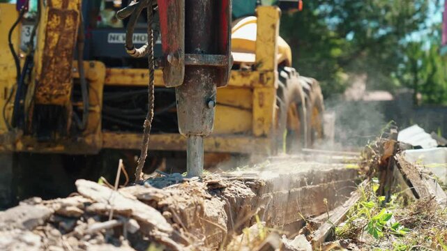 Close-up of hydraulic breaker demolishing an old concrete strip foundation beside a backhoe loader, showing dust, debris and residential site preparation.