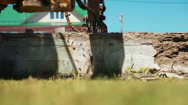 Heavy machinery with a hydraulic hammer breaking apart an old concrete wall during demolition work on a sunny day. Industrial equipment is dismantling a structure with pieces of debris falling down