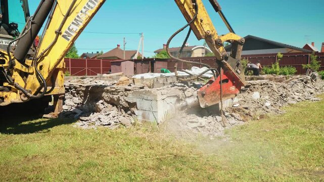 Professional yellow excavator with a hydraulic breaker attachment destroying an old concrete foundation at a construction site, creating dust and rubble during the demolition process