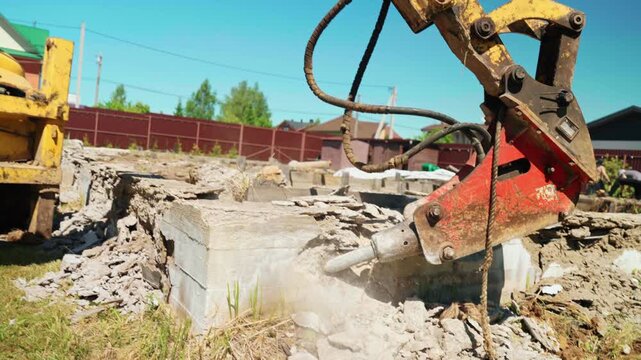 Heavy machinery with a hydraulic breaker crushing and breaking up an old concrete base at a construction site during a demolition project, with pieces of rubble and dust flying around