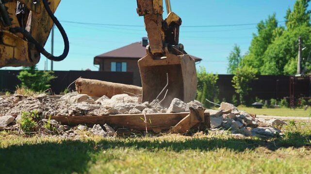 Low angle close-up of backhoe loader bucket pressing concrete rubble and broken formwork in a residential yard, showing demolition cleanup and site preparation.