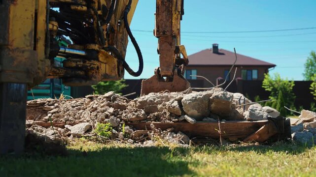 Heavy machinery with a hydraulic excavator arm breaking reinforced concrete blocks and construction waste during a demolition project on a bright, sunny day with a house in the background