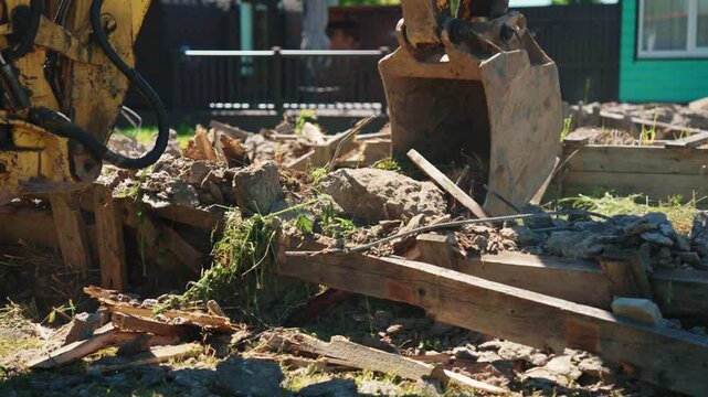 Heavy machinery with a yellow hydraulic arm and bucket digging and clearing rubble from a demolished structure. Powerful digger working on a construction site, moving earth, stones, and wood debris