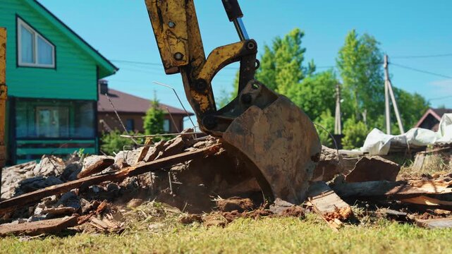Powerful yellow excavator bucket demolishing the remnants of an old building, crushing concrete, wood, and rubble on a construction site during a sunny day with a house in the background