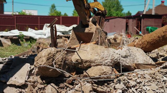Close-up of backhoe loader bucket crushing broken wood, concrete debris and exposed rebar in a residential yard, showing demolition cleanup and site preparation.