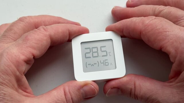 Close-up of woman's hands holding digital sensor displaying temperature and humidity, careful hands tracing display, wrinkled skin