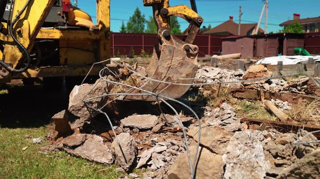 Close-up of backhoe loader bucket pulling bent rebar from broken concrete rubble in a residential yard, showing demolition cleanup and foundation removal.