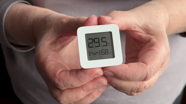 Closeup woman's hands holding digital hygrometer displaying temperature and humidity, small white square sensor, floral