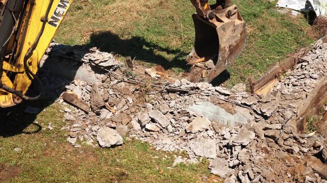 Close up top down view of a mechanical shovel bucket breaking and destroying reinforced concrete foundations during a demolition project, scattering debris and rubble over the grassy ground