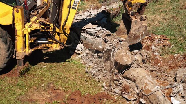 Yellow backhoe excavator's mechanical arm with a bucket digging and crushing concrete rubble during demolition work on a construction site, moving earth and destroying old foundations