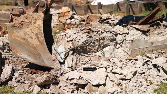 Heavy machinery excavator bucket dismantling a building's reinforced concrete foundation during demolition work at a construction site, creating a pile of debris, rubble, and rebar