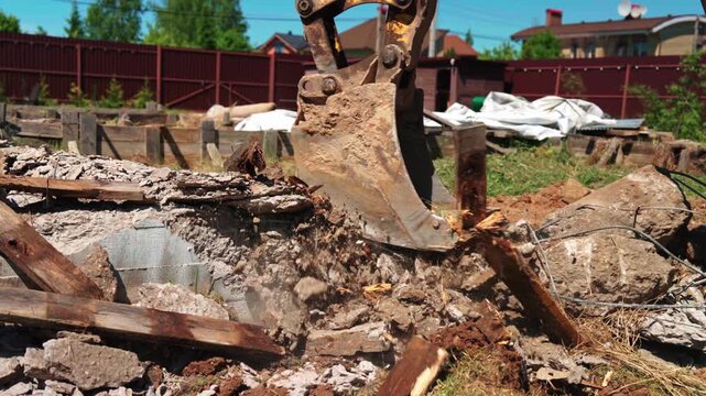 Close-up of backhoe loader bucket breaking concrete foundation debris, old wood and soil in a residential yard, showing demolition cleanup and site preparation.