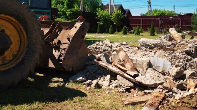 Backhoe loader bucket clearing demolished foundation debris, broken wood and exposed rebar in a residential yard, showing site cleanup and construction preparation.