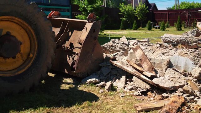 Heavy construction machinery demolishing a building foundation on a sunny day. The bulldozer bucket scoops up concrete debris and rubble from the ground during site clearance and excavation