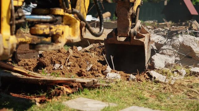 Small yellow excavator bucket digging soil and moving broken concrete rubble during demolition work on a construction site, with details of the mechanical arm and hydraulic system visible