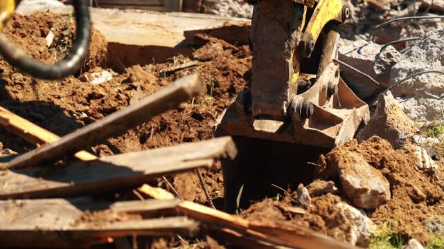 Close-up of backhoe loader bucket digging through soil, broken wood and concrete debris from an old foundation, showing demolition cleanup and site preparation.