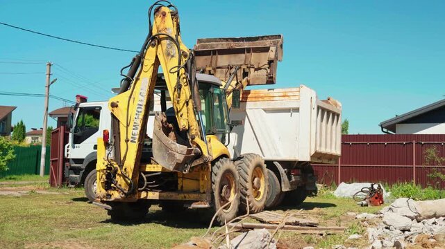 Heavy equipment working on a construction site, where a backhoe loader with a full bucket is dumping construction waste and debris into the back of a large industrial dump truck