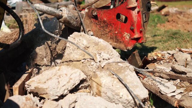 Heavy duty hydraulic hammer attached to an excavator arm breaking and crushing a large block of reinforced concrete on a construction site, creating dust and rubble during demolition work