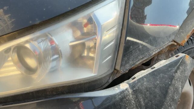 Close-up damaged black car front shows torn bumper and rusted fender, headlight illuminated, steel wheel with scuff marks, gravel