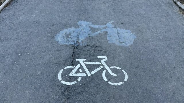 Faded bicycle lane stencil on asphalt, white bike symbol beneath old blue smudge, worn tarmac texture captured in closeup top down