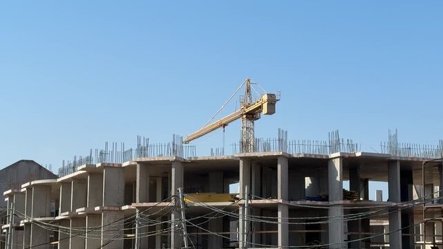 Concrete apartment building under blue sky showing raw concrete slabs and tower crane with suspended hook, scaffolding and rebar