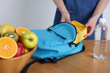 Woman putting lunch box into school bag at wooden table indoors, closeup