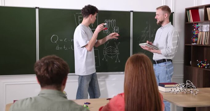 Student solving physics problems at chalkboard and teacher in classroom