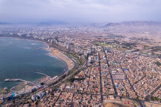 Aerial view of the Malecon de Chorrillos coastline, featuring urban sprawl, sandy beaches, fishing boats in the bay, and distant mountains in Lima, Provincia de Lima, Peru.